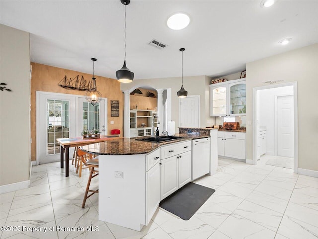 kitchen featuring sink, ornate columns, a center island with sink, white dishwasher, and dark stone counters