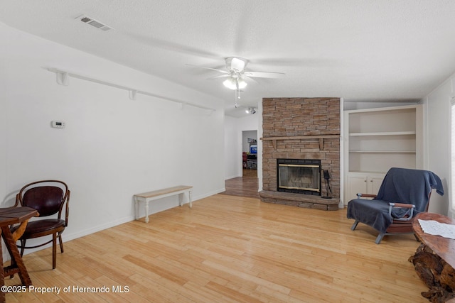 living area featuring a stone fireplace, built in features, ceiling fan, a textured ceiling, and light hardwood / wood-style flooring