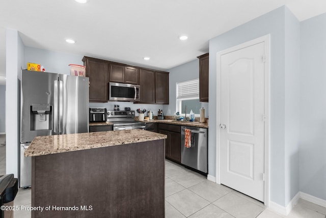 kitchen with a kitchen island, appliances with stainless steel finishes, light tile patterned floors, light stone counters, and dark brown cabinetry