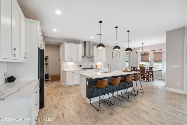 kitchen featuring wall chimney range hood, sink, a kitchen island with sink, light stone countertops, and white cabinets