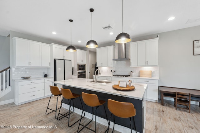 kitchen featuring sink, white cabinetry, hanging light fixtures, a center island with sink, and wall chimney range hood