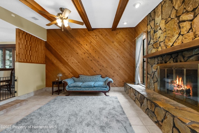 living room featuring ceiling fan, wooden walls, a fireplace, light tile patterned flooring, and beamed ceiling
