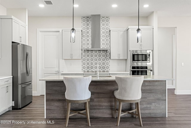 kitchen featuring wall chimney range hood, decorative light fixtures, a center island with sink, and appliances with stainless steel finishes