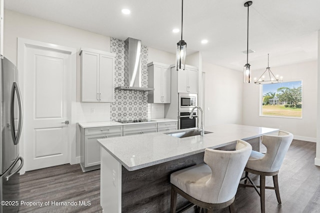 kitchen with sink, stainless steel appliances, a center island with sink, decorative light fixtures, and wall chimney exhaust hood