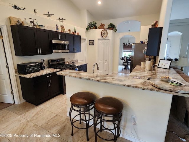 kitchen with arched walkways, appliances with stainless steel finishes, dark cabinets, a peninsula, and a kitchen bar