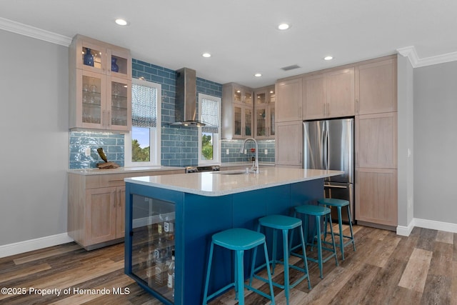 kitchen featuring a center island with sink, glass insert cabinets, freestanding refrigerator, wall chimney range hood, and backsplash