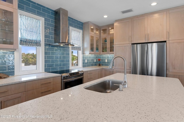 kitchen with light stone counters, stainless steel appliances, a sink, wall chimney range hood, and glass insert cabinets