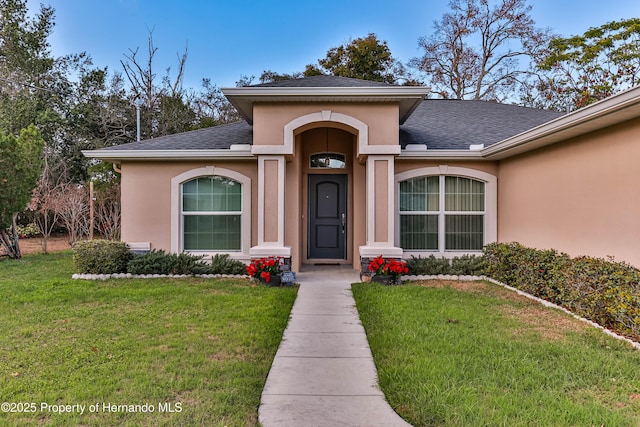 entrance to property featuring roof with shingles, a yard, and stucco siding