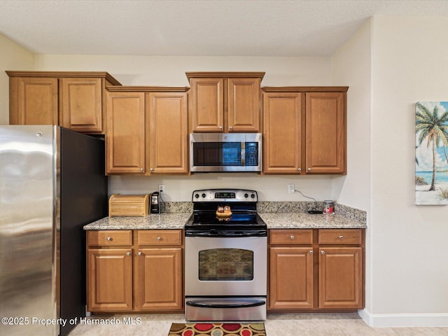 kitchen with appliances with stainless steel finishes, brown cabinetry, baseboards, and light stone countertops