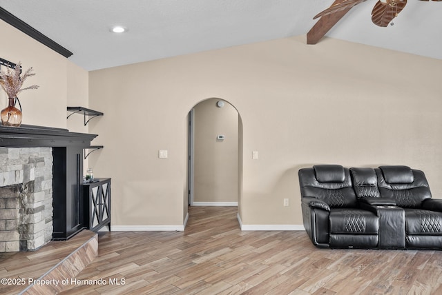 living area featuring baseboards, arched walkways, lofted ceiling with beams, a stone fireplace, and light wood-style floors