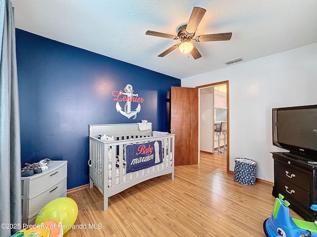 bedroom featuring a crib, visible vents, a ceiling fan, light wood-style flooring, and a textured ceiling