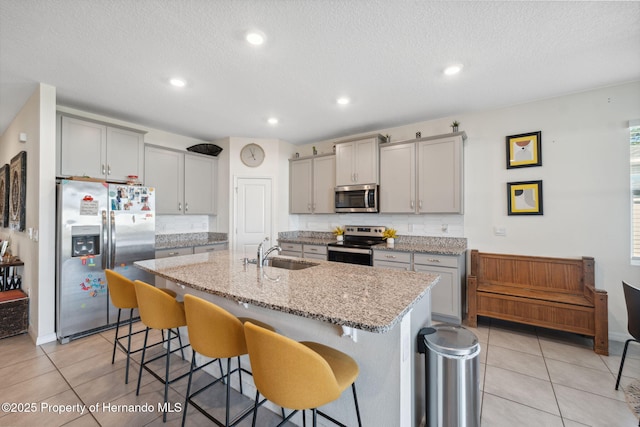 kitchen featuring a center island with sink, stainless steel appliances, gray cabinetry, a sink, and a kitchen bar