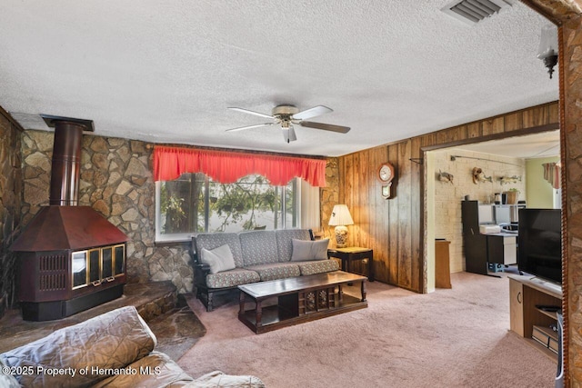 living room featuring wooden walls, carpet flooring, a ceiling fan, visible vents, and a wood stove