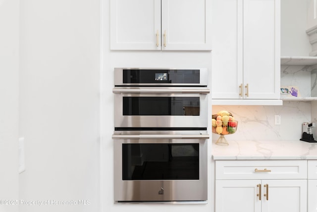 kitchen featuring light stone counters, stainless steel double oven, white cabinets, and backsplash