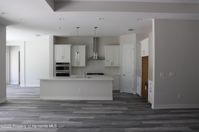 kitchen featuring white cabinets, wall chimney exhaust hood, an island with sink, a sink, and backsplash