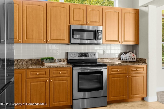 kitchen featuring light tile patterned floors, appliances with stainless steel finishes, brown cabinets, dark stone counters, and tasteful backsplash