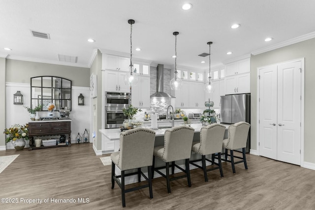 kitchen featuring visible vents, white cabinets, an island with sink, wall chimney exhaust hood, and stainless steel appliances