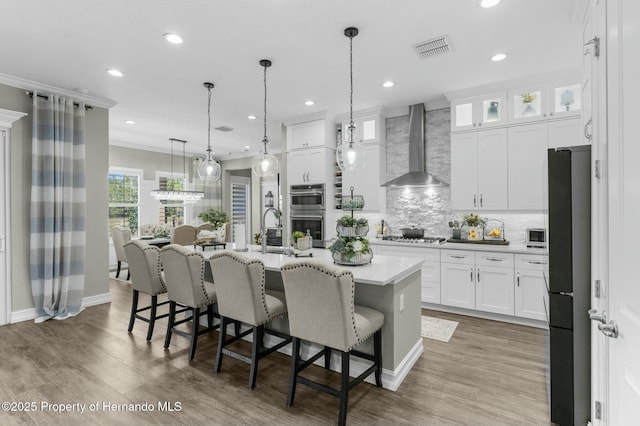 kitchen with visible vents, wall chimney exhaust hood, appliances with stainless steel finishes, a kitchen island with sink, and light countertops