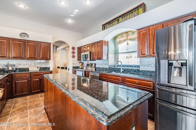 kitchen with arched walkways, a kitchen island, appliances with stainless steel finishes, dark stone countertops, and a sink