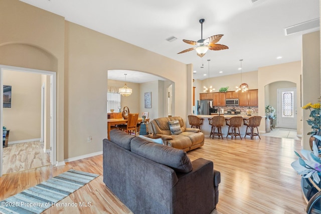 living area featuring arched walkways, visible vents, light wood-style flooring, baseboards, and ceiling fan with notable chandelier