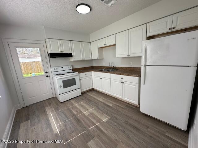 kitchen with dark countertops, dark wood-type flooring, under cabinet range hood, white appliances, and a sink