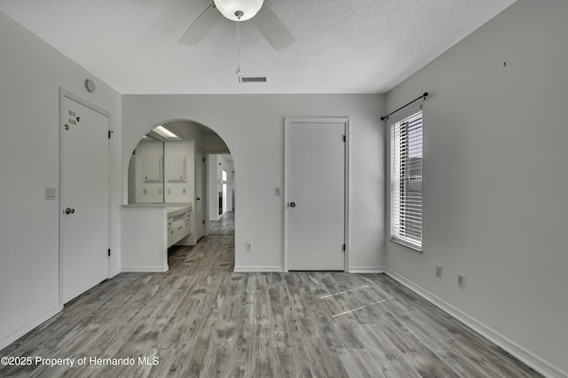unfurnished bedroom featuring a ceiling fan, arched walkways, baseboards, light wood-style flooring, and a textured ceiling