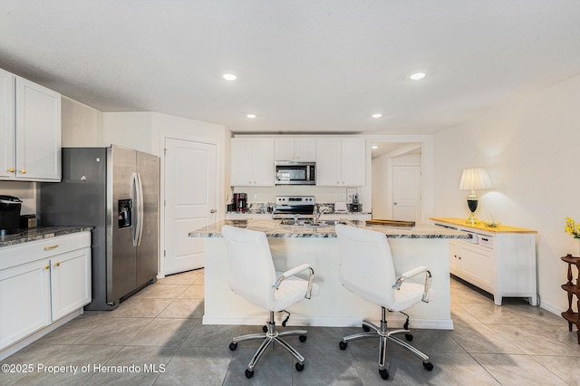 kitchen with light stone counters, light tile patterned floors, a center island with sink, appliances with stainless steel finishes, and white cabinetry