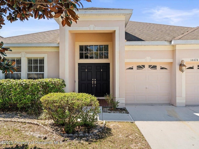 view of exterior entry with a garage, stucco siding, driveway, and a shingled roof