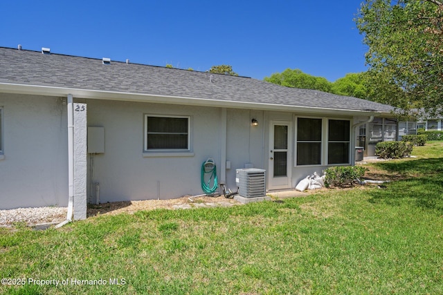 rear view of house featuring central AC unit, roof with shingles, stucco siding, and a yard