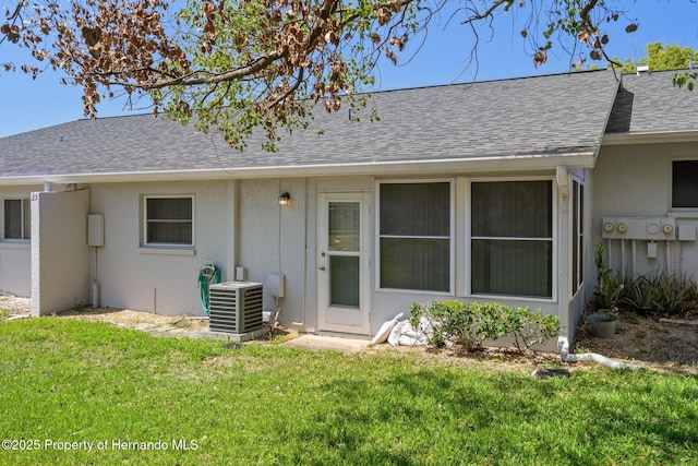 rear view of property featuring stucco siding, roof with shingles, central AC, and a yard