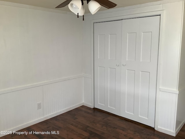 unfurnished bedroom with dark wood-style floors, a closet, a ceiling fan, and a wainscoted wall