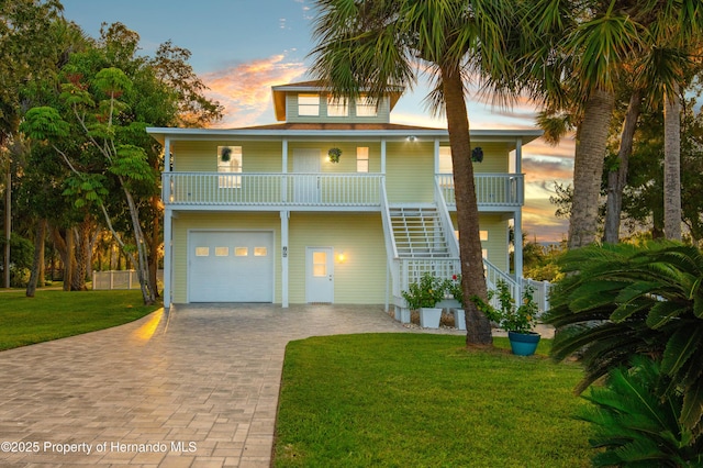 coastal inspired home featuring a garage, covered porch, decorative driveway, a lawn, and stairway