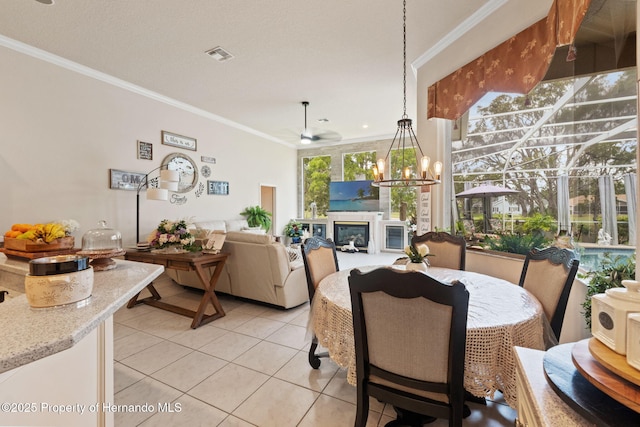 dining area featuring visible vents, ornamental molding, a glass covered fireplace, light tile patterned flooring, and ceiling fan with notable chandelier