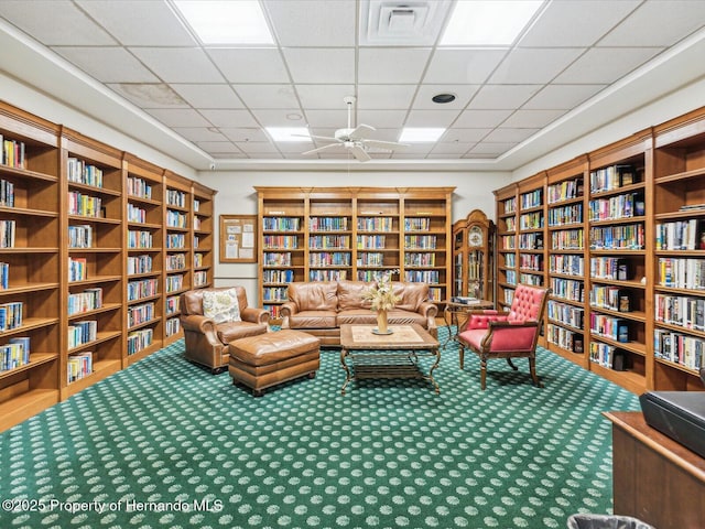 living area featuring bookshelves, carpet floors, a paneled ceiling, and ceiling fan