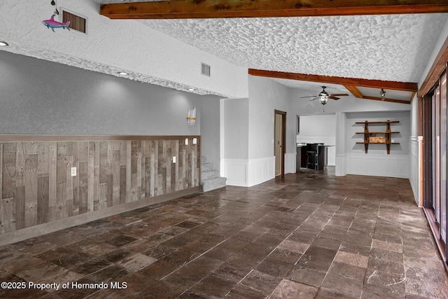 unfurnished living room featuring stairs, ceiling fan, a textured wall, wainscoting, and a textured ceiling