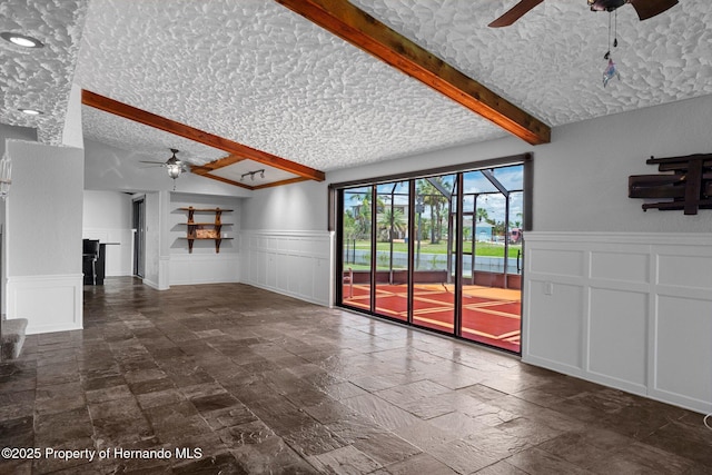 unfurnished living room with ceiling fan, wainscoting, a decorative wall, a textured ceiling, and stone tile flooring