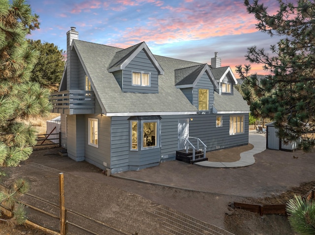 back of house at dusk featuring a chimney, a patio, a fenced backyard, roof with shingles, and a balcony