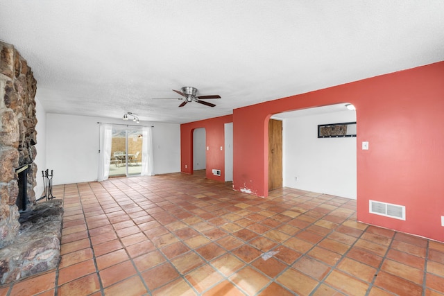 unfurnished living room with arched walkways, visible vents, ceiling fan, a stone fireplace, and a textured ceiling