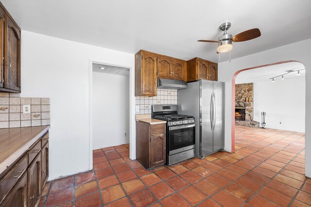 kitchen with decorative backsplash, a ceiling fan, appliances with stainless steel finishes, a stone fireplace, and under cabinet range hood