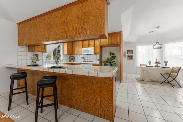 22401 Citation St, Tehachapi, CA 93561 - kitchen with white microwave, a peninsula, brown cabinets, backsplash, and light tile patterned floors