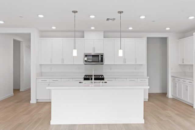 kitchen with stainless steel microwave, visible vents, an island with sink, light wood-type flooring, and light countertops