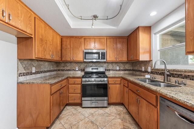 kitchen featuring light stone countertops, brown cabinets, a sink, a raised ceiling, and stainless steel appliances