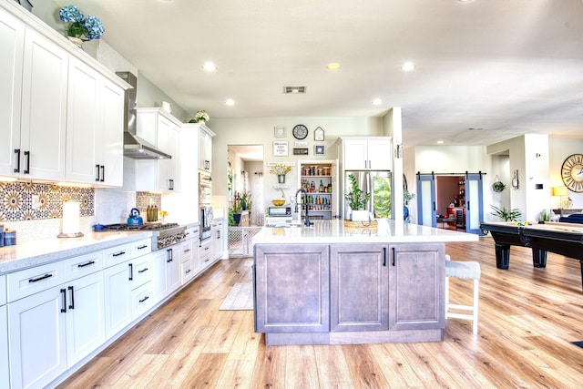 5549 Bobbie Ave, Palmdale, CA 93551 - kitchen featuring wall chimney range hood, stainless steel appliances, light wood-style flooring, an island with sink, and white cabinets
