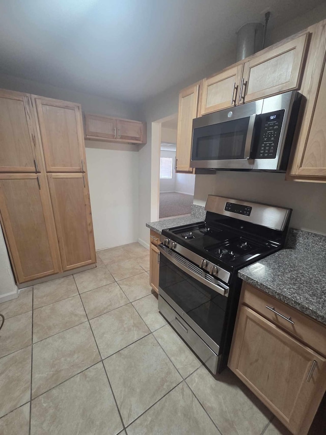 kitchen featuring appliances with stainless steel finishes, dark stone counters, light tile patterned floors, light brown cabinetry, and baseboards