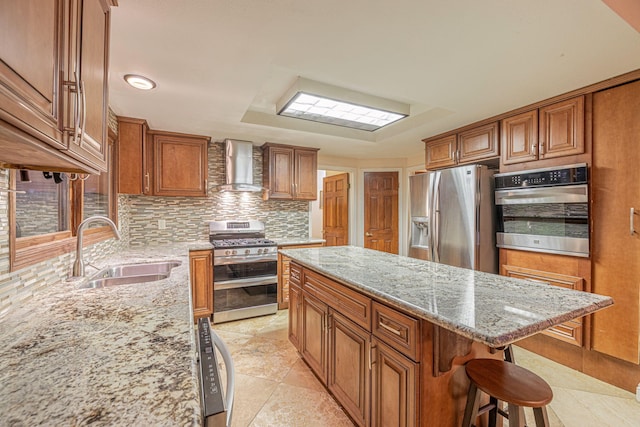 kitchen with brown cabinets, stainless steel appliances, a sink, a kitchen island, and wall chimney exhaust hood