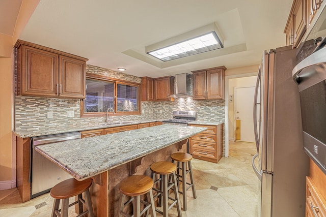 kitchen featuring a sink, a center island, appliances with stainless steel finishes, wall chimney range hood, and brown cabinets