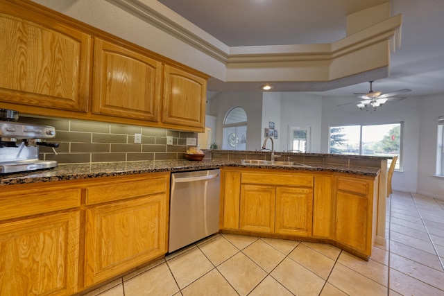 kitchen featuring stainless steel dishwasher, a sink, dark stone counters, a tray ceiling, and a peninsula