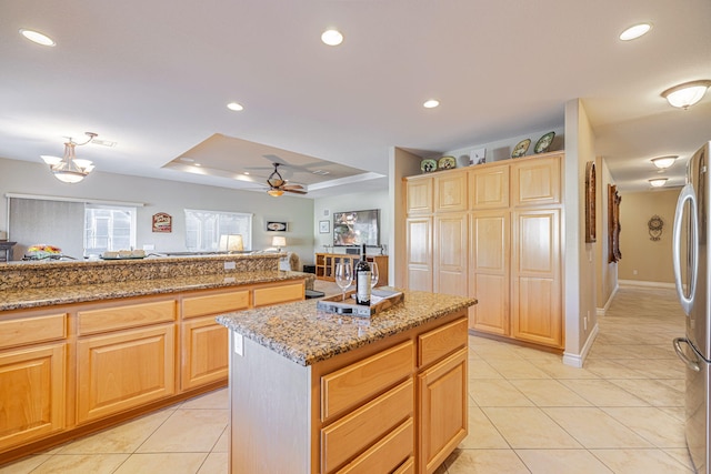 kitchen with light stone countertops, a center island, a raised ceiling, stainless steel fridge, and ceiling fan with notable chandelier