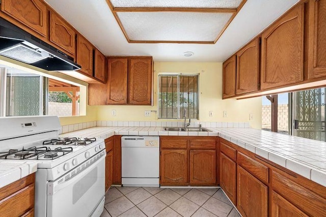 kitchen with under cabinet range hood, white appliances, a sink, tile counters, and brown cabinetry