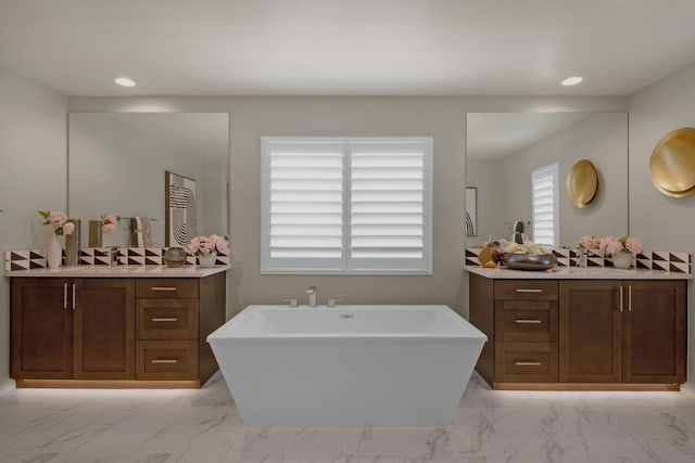 full bath featuring marble finish floor, two vanities, a freestanding tub, and recessed lighting